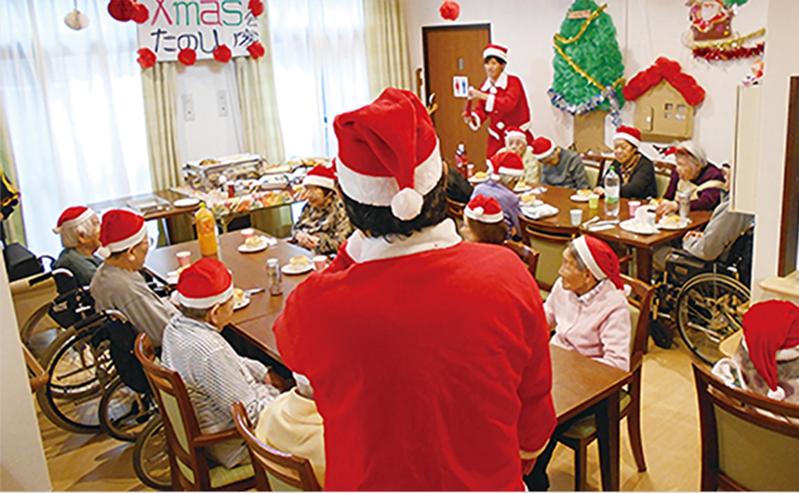 介護施設のクリスマスイベント。入居者がサンタ衣装で食卓を囲み、賑やかに楽しむ光景